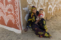 Rajasthani woman and child beside traditional painting on mud wall of house during Diwali festival, Jakholaas Kallan village, Rajasthan, India