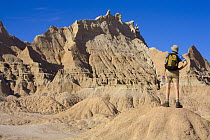 Man hiker, 52, in shorts, boots and hat, carrying backpack and water bottle, standing and looking ahead at the difficult and rocky broken terrain he wants to hike in, autumn noon, Badlands National Pa...