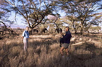 David Attenborough with sound recordist on location in Kenya, standing near elephant herd, about to do a piece to camera