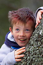 Young boy peering around Birch tree, Cairngorms, Scotland, UK Model released.