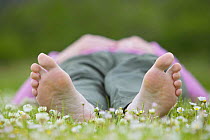 Woman lying barefoot in field of daisies, enjoying nature on a summer day, Cairngorms National Park, Scotland, UK. Soles of feet visible Model released.