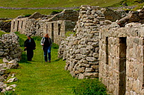 People walking past abandoned stone brick houses, Hirta, Saint / St Kilda  Island, Western Islands, Outer Hebrides, Scotland.
