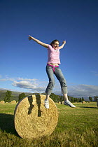 Girl jumping off large round hay bale, Scotland, UK Model released.