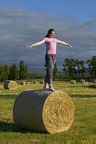 Girl balancing on large round hay bale, Scotland, UK Model released.