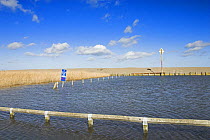 Coastal car park flooded by water from freshwater marsh, sea wall in background, North Norfolk, UK
