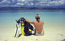 Woman sitting on beach with scuba tank and fins, looking out to sea, Dimakya Island, Palawan, Philippines