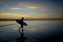 Surfer carrying surfboard up beach silhouetted at sunset, Sandymouth bay, Cornwall, UK