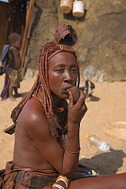 Woman of the Himba tribe, Kaokoland, wearing distinctive headdress and clothing, smoking a pipe outside a traditional mud and dung hut. Namibia 2007