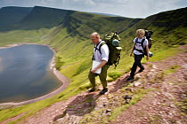 Walkers carrying backpacks hiking above Llyn y Fan Fach, a glacial cirque, beneath Black Mountain in the Brecon Beacons National Park, Powys, Wales
