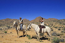 Two people horse riding in the Namib desert, one person is looking through binoculars, Namibia