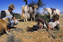 Two people with horses (Equus caballus) and a dog, cooking and taking a rest stop from riding, Namib desert, Namibia