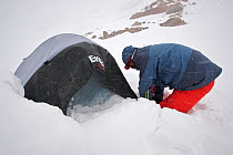 Skier Phil Atkinson digging out his tent after an all night blizzard at camp site in the upper Sky Top Lakes Valley near Granite Peak, Beartooth Mountains, Montana, USA.  May 2008
