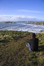 Woman sitting beside coastal path near Bantham Bay, with Burgh Island in the distance. South Devon, UK. January 2009.
