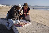 Traveller sitting on wall, haggling for local cakes on Essaouira beach, November 2008.