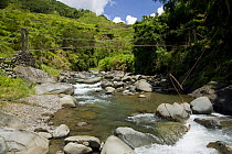 River and bridge in Banaue Rice Terraces, Philippines. In the foreground, a device can be seen that uses water to pull a long line going up to the rice fields to wave a the flag that frightens away bi...