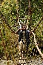 Boy crossing a pole and vine suspension bridge over river carrying a machette and photographer's tripod bag, Crater Mountain Wildlife Management Area, Eastern Highlands Province, Papua New Guinea, Sep...