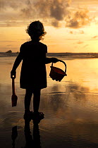 Little girl with bucket and spade silhouetted against setting sun on Summerleaze beach, Bude, North Cornwall, UK. July 2009, Model released.