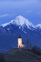 Church of St. Primoz at dusk with the peak of Storzic in the Kamnik-Savinja Alps in the background, Jamnik, Gorenjska, Slovenia, Europe, March 2009