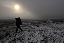 Photographer, Vincent Munier, walking with rucksack through snow covered landscape with sun shining through storm sky, Forollhogna National Park, Norway. On location for Wild Wonders of Europe, Septem...