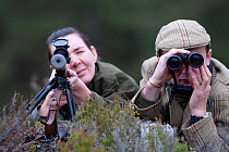 Deer stalker using binoculars to spot deer for guest looking down telescopic sight of gun, Glenfeshie, Cairngorms, Scotland, UK, June 2008, model released