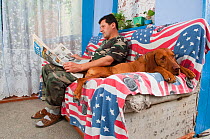 Man sitting on sofa reading newspaper next to a large dog, Moldova, June 2009