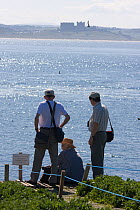 Three people watching Puffin (Fratercula arctica) colony on Farne Islands, with Bambergh Castle in the distance, Northumberland, UK, June 2009