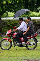 Two men on a motorcycle using an umbrella to protect them from the monsooon rain, Bangladesh, November 2008