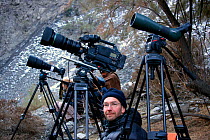 Cameraman, Mark Smith, waiting patiently for a sight of the elusive Snow leopard, with a range of binoculars, telescopes and cameras all pointed towards a cave the snow leopard is thought to use, for...