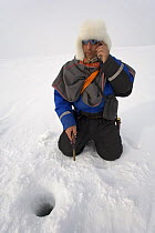 Saami reindeer herder talking on his mobile phone while ice fishing for Arctic Char on a lake near his winter pastures. Kautokeino, Finnmark, North Norway, March 2007.