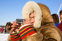 Nenets spectator using his mobile phone to photograph a reindeer race at a herders festival in the Yamal. Western Siberia, Russia, February 2009. Editorial use only.