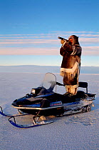 Inuit hunter standing on his snowmobile to scan the surrounding ice for seals. Igloolik, Nunavut, Canada, 1995.