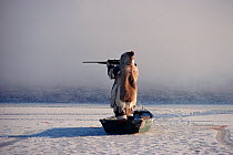 Inuk standing in floe edge boat at the ice edge, aiming his rifle while hunting. Igloolik, Nunavut, Canada, 1990
