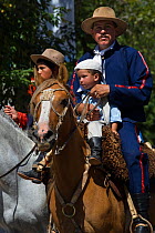 Two generations of traditionally dressed gauchos (cowboys) ride a horse (Equus caballus) during the parade of the Fiesta De La Patria Gaucha, Tacuarembo, Uruguay, April 2008