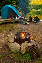Camp fire burning at waterfront campsite in Lake Francis State Park. Couple relaxing in chairs overlooking lake.  Pittsburg, New Hampshire, USA, August 2008.