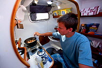 Young man cooking below decks aboard 30ft Tiki catamaran "Abaco". Exumas, Bahamas, Caribbean. June 2009, Model released.