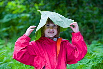 Young girl smiling, with large Butterbur (Petasites sp) leaf on her head, Scotland, UK, May 2009. Model released