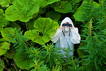 Boy in protective white suit and mask in the wilds surrounded by green vegetation, using binoculars, Scotland, UK, Model released