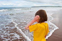 Little girl with shell pressed to her ear, listening to the sea, on the beach, Scotland, UK. Model released