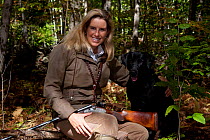 Lady hunter with .12 gauge shotgun and black Labrador Retriever in autumn woodland, sitting on log; Putnam, Connecticut, USA. Model Released