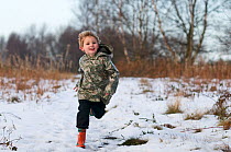 Young boy in red wellies running through snow in winter, Norfolk, England, UK. Model released