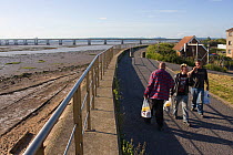 Group of people walking along Severnside footpath with Severn Bridge in background. This area of the Severn estuary could be potentially threatened by proposed tidal barrage. Gloucestershire, England,...