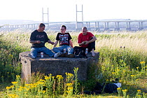 Three men sitting on wartime defences, with Severn Bridge behind. This area of the Severn estuary many be threatened by proposed tidal barrage. Gloucestershire, England, UK April 2010