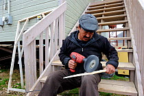 Inuit man sitting on steps, carving a Narwhal tusk (Monodon monoceros) with an electronic power tool, Qikiqtarjuaq village, Baffin Island, Nunavut, Canada, August 2010