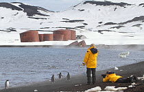 Tourists photographing Gentoo penguins (Pygoscelis papua) at Whalers Bay, with the abandoned whaling station beyond. Deception Island, South Shetland Islands, Antarctica, 2009.