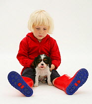 Young blonde haired girl, sitting with King Charles Spaniel puppy and welly boots. Model released