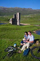 Couple relaxing during bike ride in front of Ardvreck Castle. Loch Assynt, Sutherland, Scotland, July Model released