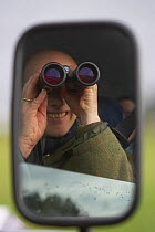 Man reflected in a wing mirror, looking from the window of a car with binoculars. Islay, Scotland, March.