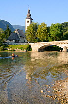 Man paddles kayak towards Ribcev Laz church and bridge at the south end of Lake Bohinj, with the forested Julian Alps in the background, Triglav National Park, Slovenia, July 2010.