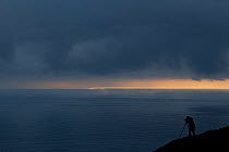 Silhouette of photographer Mark Hamblin against dramatic light over sea at dusk, Hermaness NNR, Unst, Shetland Islands, Scotland, UK, June 2010.  Model released.