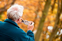 Elderly man taking digital photographs of Beech woodland in autumn, Beacon Hill Country Park, The National Forest, Leicestershire, UK, November 2010. Model released